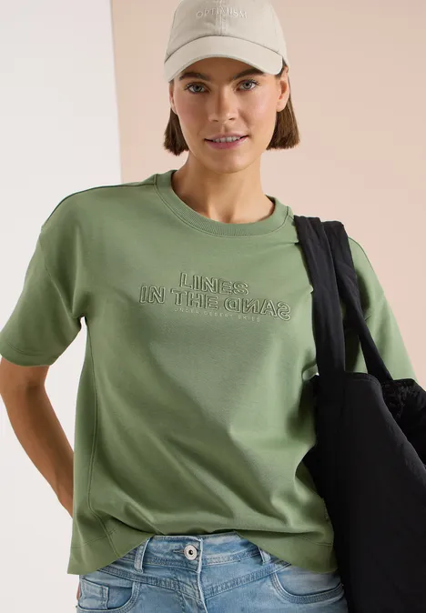 Person in hellgrünem T-Shirt, Jeans, Beanie und schwarzer Tasche vor neutralem Hintergrund